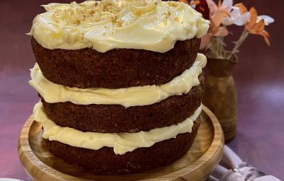 Foto em tons de marrom da receita de carrot cake servida sobre uma tábua redonda de madeira. Ao lado esquerdo há um prato de porcelana com um garfo prateado, ao lado direito há um lenço cor de rosa