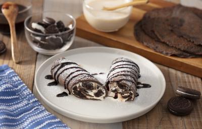 Foto da receita de Burrito Doce de Negresco, decorado com calda de chocolate e açúcar, em um prato branco grande, sobre uma bancada de madeira decorada com um tecido azul, alguns Biscoitos Negresco e dois poptinhos com caldas de chocolate