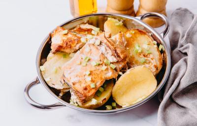 Fotografia de postas de peixe assado com molho à base de vinho e batatas. Por cima, cebolinha salpicada, o assado está dentro de um recipiente de alumínio fundo, que está sobre uma mesa de mármore ao lado de uma toalha na cor cinza.