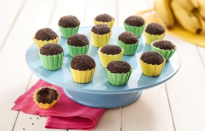 Fotografia em tons de marrom em uma bancada de madeira de cor branca. Ao centro, uma boleira azul contendo os brigadeiros. Ao lado, há um pano rosa com um brigadeiro em cima. Ao fundo, há algumas bananas.