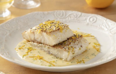 Filés de bacalhau servidos com molho cremoso de ervas e raspas de limão, apresentados em prato branco elegante.