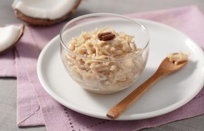 Fotografia em tons de branco e rosa de uma bancada branca com um paninho rosa, sobre ela um prato redondo branco com um recipiente de vidro com cocada e uma colher. Ao fundo um coco.