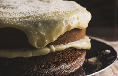 Fotografia de um bolo de abóbora com creme de coco por cima. O bolo está recheado com o mesmo creme, e está sobre um prato raso preto. Fotografia de um bolo de abóbora com creme de coco por cima. O bolo está recheado com o mesmo creme, e está sobre um prato raso preto.