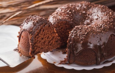 Bolo de chocolate fofinho em forma de bundt, coberto com uma camada brilhante de chocolate cremosa que escorre pelas laterais. Decorado com raspas de chocolate, o bolo é apresentado em um fundo de madeira quente, sugerindo um toque caseiro Bolo de chocolate fofinho em forma de bundt, coberto com uma camada brilhante de chocolate cremosa que escorre pelas laterais. Decorado com raspas de chocolate, o bolo é apresentado em um fundo de madeira quente, sugerindo um toque caseiro