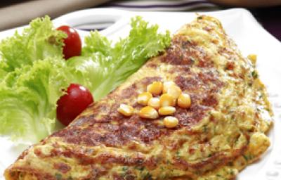 Fotografia em tons de verde em uma mesa com uma toalha listrada com marrom e bege, um prato branco redondo com uma omelete com milho e salada de alface e tomate cereja ao lado do prato.