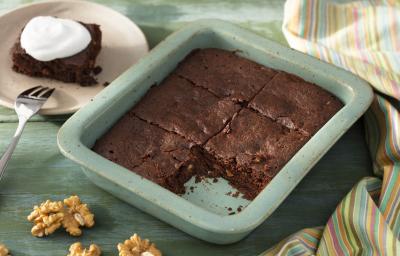 Fotografia em tons de verde em uma bancada de madeira verde, um recipiente verde quadrado com o brownie de chocolate com nozes e um pano colorido ao lado. Ao fundo, um prato redondo bege com uma fatia do brownie. Fotografia em tons de verde em uma bancada de madeira verde, um recipiente verde quadrado com o brownie de chocolate com nozes e um pano colorido ao lado. Ao fundo, um prato redondo bege com uma fatia do brownie.