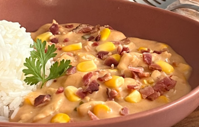 A iumagem mostra um prato com uma porção de frango cremoso com pedaços de bacon e milho e uma porção de arroz ao lado. O prato é marrom avermelhado e está apoiado em uma tábua de madeira com um guardanapo listrado e um garfo ao fundo.