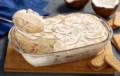 Fotografia em tons de branco, com tábua de madeira com um recipiente de vidro alto com o pavê de coco com biscoitos maisena São Luiz espalhados pela mesa. Fotografia em tons de branco, com tábua de madeira com um recipiente de vidro alto com o pavê de coco com biscoitos maisena São Luiz espalhados pela mesa.