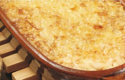 Fotografia em tons de marrom em uma bancada com um suporte para panela de madeira, uma travessa de barro com as cebolas gratinadas dentro dela e em cima do suporte. Ao lado, potinho com queijo ralado. Fotografia em tons de marrom em uma bancada com um suporte para panela de madeira, uma travessa de barro com as cebolas gratinadas dentro dela e em cima do suporte. Ao lado, potinho com queijo ralado.