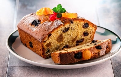 Fotografia de uma fatia de bolo de frutas em um prato, com uma crosta dourada e interior úmido, repleto de frutas secas. O bolo é decorado com açúcar de confeiteiro, folhas de hortelã e frutas cristalizadas.