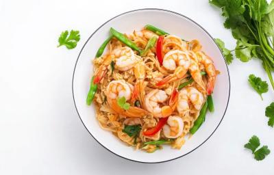 Fotografia vista de cima em tons claros de uma mesa branca. Sobre a mesa tem um prato fundo branco ao lado de um maço de coentro. Dentro do prato tem uma receita de pad thai com camarões.