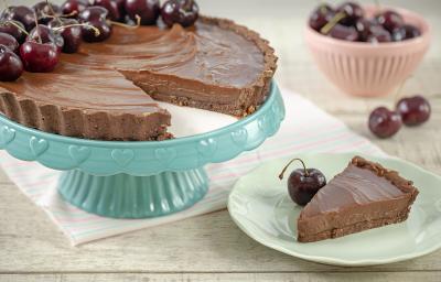 Fotografia em tons de marrom, branco, azul e rosa de uma bancada cinza vista de frente. Um recipiente azul contém uma torta de chocolate com varias cerejas e com uma fatia retirada, ao lado um prato branco com a fatia de torta e uma cereja do lado.