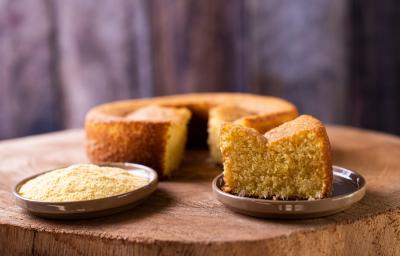 Foto da receita de bolo de milho de liquidificador, redondo, dourado, com uma fatia mais à frente em um prato e ao lado um punhado de fubá em outro prato. Ao fundo é possível ver o bolo inteiro Foto da receita de bolo de milho de liquidificador, redondo, dourado, com uma fatia mais à frente em um prato e ao lado um punhado de fubá em outro prato. Ao fundo é possível ver o bolo inteiro