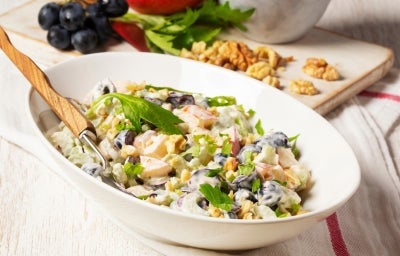 Fotografia de uma salada em uma tigela oval branca, com maçãs, melão e uvas pretas em um molho cremoso, decorada com nozes e salsa, acompanhada de um garfo, em um ambiente rústico com ingredientes frescos ao fundo.