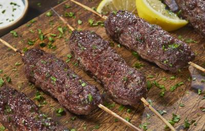 Fotografia de quatro espetinho de carne moída sobre uma tábua de madeira, e por cima, salsinha salpicada. Ao fundo, um molho de pepino dentro de um recipiente de fundo e pequeno, e ao lado, dois pedaços de limão siciliano. Fotografia de quatro espetinho de carne moída sobre uma tábua de madeira, e por cima, salsinha salpicada. Ao fundo, um molho de pepino dentro de um recipiente de fundo e pequeno, e ao lado, dois pedaços de limão siciliano.