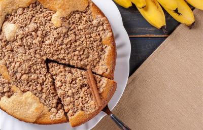 Fotografia vista de cima de uma cuca de banana com farofa sobre um prato de vidro branco raso, apoiado em uma mesa de madeira preta com um pano marrom de tom claro. Fotografia vista de cima de uma cuca de banana com farofa sobre um prato de vidro branco raso, apoiado em uma mesa de madeira preta com um pano marrom de tom claro.