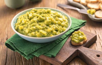 Foto da receita de Guacamole. Observa-se um pote redondo com a guacamole, sobre um guardanapo verde e uma tábua de madeira. Ao lado do recipiente, uma torrada redonda com o guacamole. As mesmas torradas decoram o fundo da foto.