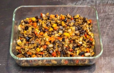 Em cima de uma mesa, refratário com berinjelas, pimentões e cebolas picados, regados com azeite.
