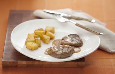 Fotografia em tons de branco e marrom de uma mesa de madeira, vista de lado, ao centro um prato redondo branco, com o filé regado com molho e batatas assadas para acompanhar. Ao lado talheres e um guardanapo de linho em tons beges.