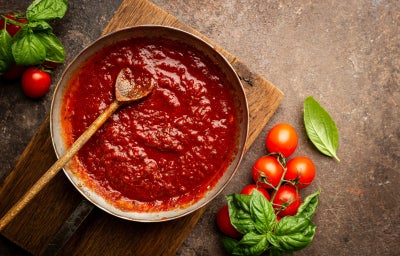 Imagem mostra um recipeinte com molho de tomate vermelho e uma colher dentro, folhas de manjericão e tomates ao lado