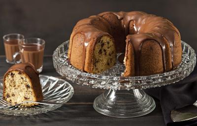 Fotografia em tons de marrom e preto de uma bancada preta vista de frente. Contém um prato redondo transparente com uma fatia de bolo com calda de chocolate por cima, um recipiente transparente para servir o bolo com o bolo sem uma fatia e calda por cima. Fotografia em tons de marrom e preto de uma bancada preta vista de frente. Contém um prato redondo transparente com uma fatia de bolo com calda de chocolate por cima, um recipiente transparente para servir o bolo com o bolo sem uma fatia e calda por cima.