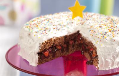 Fotografia em tons de rosa em uma bancada cinza com um suporte para bolo central na cor rosa com o bolo de aniversário confeitado em cima com cobertura branca e confeitos coloridos, cortado ao meio e recheado com chocolate e morango. Fotografia em tons de rosa em uma bancada cinza com um suporte para bolo central na cor rosa com o bolo de aniversário confeitado em cima com cobertura branca e confeitos coloridos, cortado ao meio e recheado com chocolate e morango.