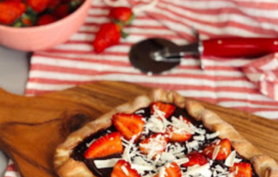 Fotografia em tons de vermelho, em uma bancada de madeira de cor branca. Ao centro, um pano listrado vermelho contendo uma tábua de madeira de cor marrom e em cima está a pizza. Ao fundo, um recipiente com morangos e alguns espalhados no chão. Fotografia em tons de vermelho, em uma bancada de madeira de cor branca. Ao centro, um pano listrado vermelho contendo uma tábua de madeira de cor marrom e em cima está a pizza. Ao fundo, um recipiente com morangos e alguns espalhados no chão.