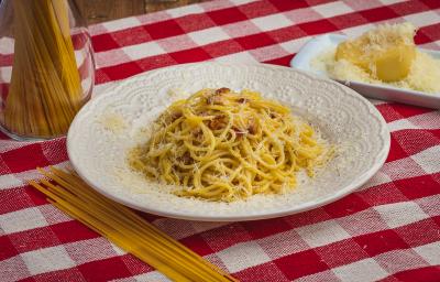 Foto de uma mesa com uma toalha xadrez vermelha e branca. Sobre ela há um prato branco com a receita de Spaghetti à Carbonara, alguns macarrões crus espalhados e num pote e, do lado direito, um pedaço de queijo com queijo ralado. Foto de uma mesa com uma toalha xadrez vermelha e branca. Sobre ela há um prato branco com a receita de Spaghetti à Carbonara, alguns macarrões crus espalhados e num pote e, do lado direito, um pedaço de queijo com queijo ralado.