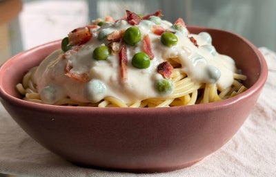 Fotografia mostra um macarrão com molho branco, ervilhas e pedaços de bacon em um bowl.