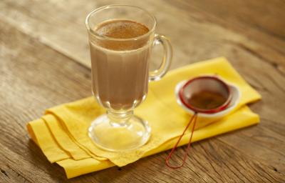Fotografia em tons de marrom e amarelo de uma bancada de madeira com um paninho amarelo, sobre ele uma xícara transparente com leite, chocolate baton derretido e canela em pó. Ao lado uma peneira vermelha com canela. Fotografia em tons de marrom e amarelo de uma bancada de madeira com um paninho amarelo, sobre ele uma xícara transparente com leite, chocolate baton derretido e canela em pó. Ao lado uma peneira vermelha com canela.