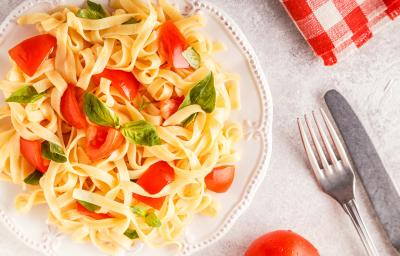 fotografia em tons de branco, amarelo e vermelho de uma bancada branca vista de cima. Contém um prato redondo branco com macarrão tipo talharim com pedaços de tomate e folhas de manjericão. Ao lado garfo e faca para servir. fotografia em tons de branco, amarelo e vermelho de uma bancada branca vista de cima. Contém um prato redondo branco com macarrão tipo talharim com pedaços de tomate e folhas de manjericão. Ao lado garfo e faca para servir.