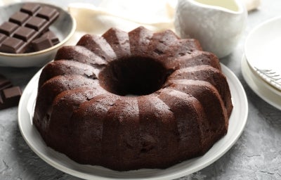 Bolo de chocolate em forma de bundt, com cor marrom escura e superfície brilhante, servido em um prato branco. Ao fundo, um jarro de cerâmica branca e pedaços de chocolate.
