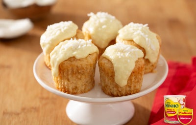 Bolinho de coco com cobertura clara e coco ralado, dispostos sobre prato com pedestal, em mesa de madeira.