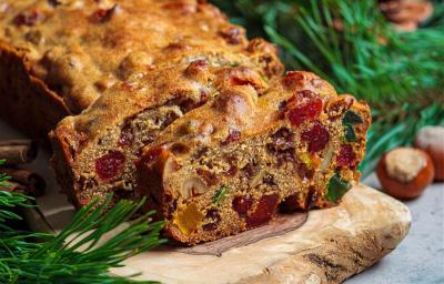 Fotografia de um bolo de frutas cristalizadas e secas com whisky e especiarias, feito na forma pequena de bolo inglês. Tem duas fatias cortadas na frente do restante do bolo, que está sobre um apoio de madeira, ao lado de paus de canela.