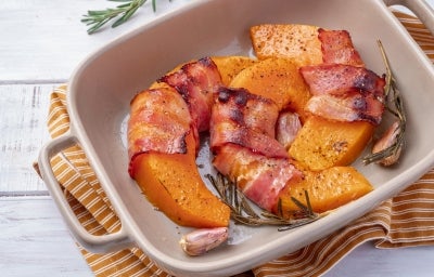 Em cima de uma mesa, há um refratário quadrado com abóboras cortadas em meia-lua enroladas com bacon.