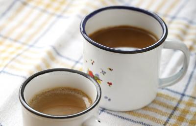 Fotografia em tons de branco, bege e azul, com duas canecas brancas com borda azul contendo bebida em tons café-com-leite, sobre guardanapo branco com listras azul e bege, tudo sobre bancada de madeira.