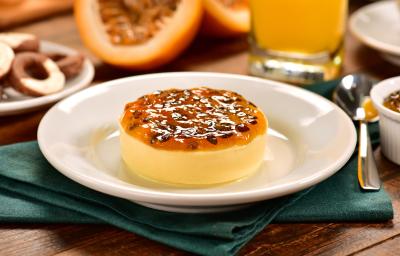 Fotografia de um flan de maracujá com calda por cima, sobre um prato branco e raso branco. Ao fundo, um maracujá cortado na metade, uma jarra com o suco da fruta e alguns recipientes brancos de cerâmica sobre uma mesa de madeira.