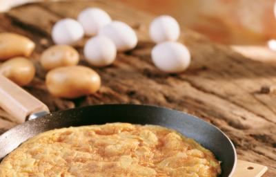 Fotografia em tons de bege em uma bancada de madeira, uma frigideira preta com uma fritada de batata dentro. No fundo da foto existem alguns ovos.