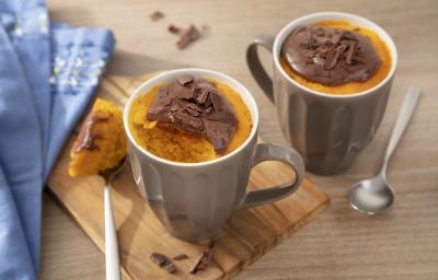 Fotografia em tons de laranja em uma bancada de madeira clara, uma tábua de madeira, um paninho azul, duas canecas marrons com o bolo de cenoura dentro de cada uma e por cima o brigadeiro de Nescau. Fotografia em tons de laranja em uma bancada de madeira clara, uma tábua de madeira, um paninho azul, duas canecas marrons com o bolo de cenoura dentro de cada uma e por cima o brigadeiro de Nescau.
