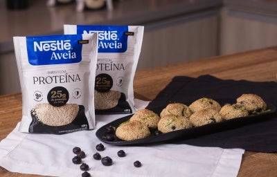 Foto da receita de bolinho de limão e mirtilo com aveia servida em diversas porções sobre uma louça preta em cima de uma mesa de madeira com duas embalagens de nestlé aveia proteína ao lado