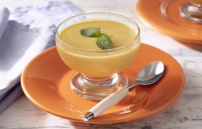 Fotografia em tons de amarelo em uma bancada de madeira de cor branca. Ao centro, um prato laranja com uma taça em cima contendo a mousse e uma colher ao lado. Ao fundo, há um prato laranja e um pano lilás.