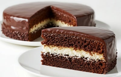 Fotografia de um bolo de chocolate redondo, coberto com ganache suave. Uma fatia revela camadas de chocolate fofinho e um recheio branco de coco.