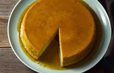 Foto da Receita de Pudim. Observa-se um pudim com um pedaço faltante em um prato grande redondo de cerâmica sobre uma mesa de madeira escura.