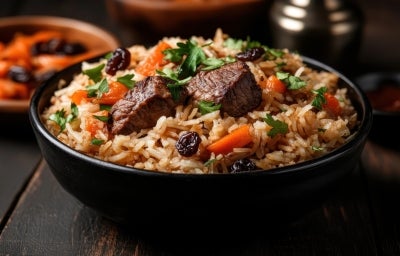 Em um cima de uma mesa, tigela redondo com arroz, carne de boi picada, cenoura e uvas-passas, decorado com cebolinha picada.