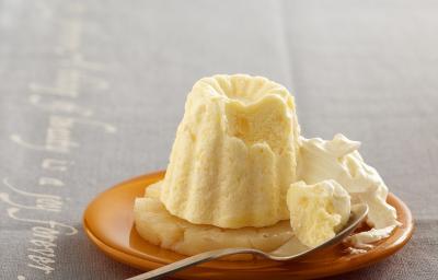Fotografia em tons de branco em uma bancada de madeira de cor cinza. Ao centro, um prato marrom contendo o flan com uma colher ao lado. Fotografia em tons de branco em uma bancada de madeira de cor cinza. Ao centro, um prato marrom contendo o flan com uma colher ao lado.