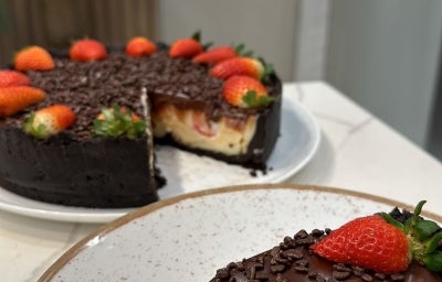 Fotografia da torta quase inteira sobre um prato raso branco, apoiado em uma mesa branca. Na frente da foto tem um prato de vidro grande com uma fatia da torta com ganache.
