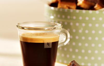 fotografia em tons de marrom, bege e verde tirada de frente de uma xicara transparente com café, e ao fundo um recipiente verde com bolinhas brancas que contém o Caramel Shortcake