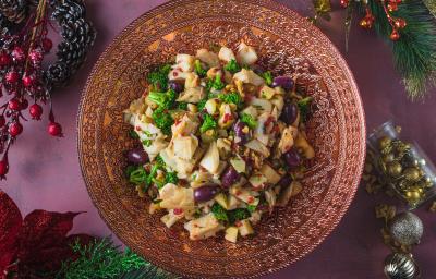 Foto vista de cima da receita de salada de bacalhau em uma travessa redonda vermelha, sobre uma mesa em tom vermelho toda decorada com motivos natalinos ao redor