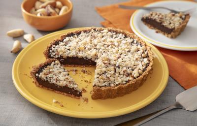 Fotografia em tons de amarelo em uma bancada de madeira de cor cinza. Ao centro, um prato amarelo contendo o cheesecake com uma espátula ao lado. Ao fundo, há um pano laranja com um pires branco em cima contendo uma fatia da sobremesa e algumas castanhas. Fotografia em tons de amarelo em uma bancada de madeira de cor cinza. Ao centro, um prato amarelo contendo o cheesecake com uma espátula ao lado. Ao fundo, há um pano laranja com um pires branco em cima contendo uma fatia da sobremesa e algumas castanhas.