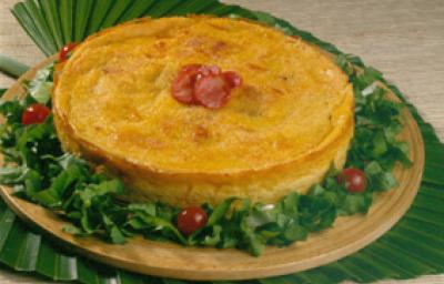 Fotografia em tons de amarelo em uma bancada de madeira clara, uma toalha verde em formato de folha, uma tábua de madeira redonda com a torta caipira em cima dela e decorada com salada verde e tomates-cereja. Fotografia em tons de amarelo em uma bancada de madeira clara, uma toalha verde em formato de folha, uma tábua de madeira redonda com a torta caipira em cima dela e decorada com salada verde e tomates-cereja.